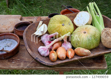 Rustic wooden bowl filled with vegetables including garlic, onions, squash, and celeriac arranged on a wooden table outdoors. A bowl of creamy soup and rustic kitchen tools add to the rural setting Rustic wooden bowl filled with vegetables including garlic, onions, squash, and celeriac arranged on a wooden table outdoors. A bowl of creamy soup and rustic kitchen tools add to the rural setting 127499059