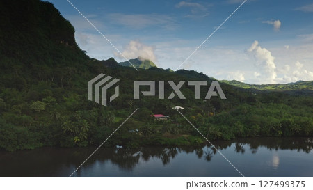 Verdant jungle landscape with a mountain in the background, reflecting in a serene river. A small house with a red roof nestled among dense greenery under a clear blue sky Verdant jungle landscape with a mountain in the background, reflecting in a serene river. A small house with a red roof nestled among dense greenery under a clear blue sky 127499375