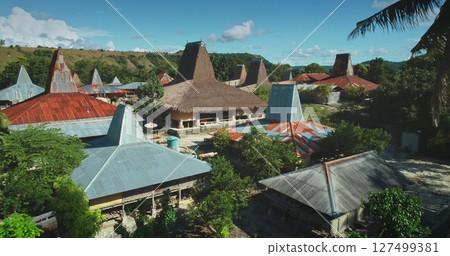 Aerial view of a village in Sumba, Indonesia, showing traditional houses with conical roofs made of straw and metal sheets, surrounded by lush vegetation under a clear blue sky 127499381