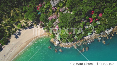 Turquoise water gently wash sand beach and rocky coast, lush tropical vegetation and charming bungalows in Thailand idyllic paradise. Exotic travel vacation getaway. Drone flight. Aerial top down view Turquoise water gently wash sand beach and rocky coast, lush tropical vegetation and charming bungalows in Thailand idyllic paradise. Exotic travel vacation getaway. Drone flight. Aerial top down view 127499408