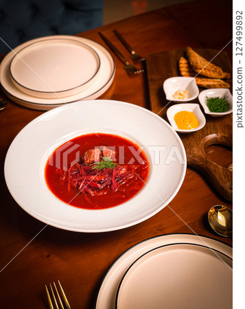 Traditional Ukrainian borscht with beef and dill served in a white bowl, styled with condiments and pastry on a warm wooden restaurant table 127499892