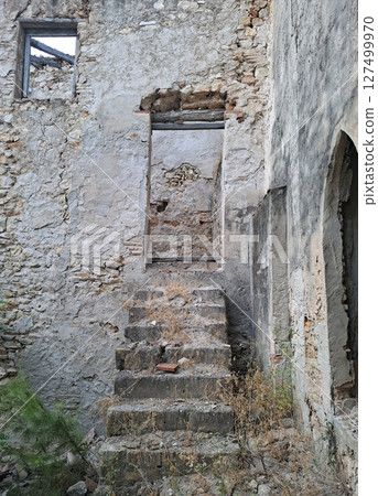 Ruins of a medieval house in Alicante reveal crumbling stone steps leading to a weathered doorway. Nature reclaims the structure, highlighting its eerie and abandoned atmosphere. 127499970