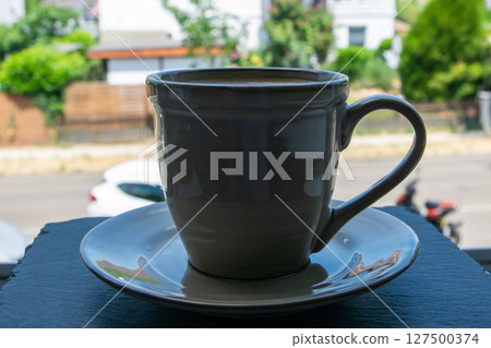 Green cup of coffee stay on the black stone window sill with a beautiful view of the summer street.  127500374