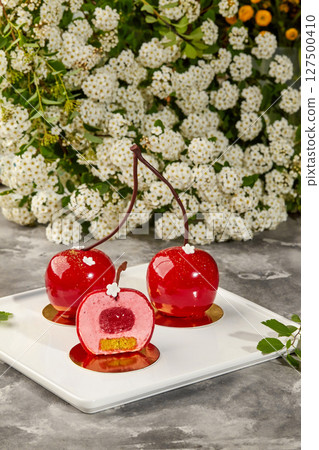 Cherry-shaped mousse cakes with mirror glaze on white plate displayed near white flowers Cherry-shaped mousse cakes with mirror glaze on white plate displayed near white flowers 127500410