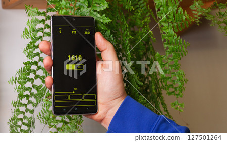 Hand holding smartphone displaying data in a lush green plant environment for sustainability efforts 127501264