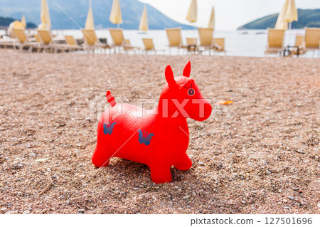 Red inflatable toy on a pebble beach with sun loungers and sea view. Childhood joy, summer vacation, and beachside relaxation. 127501696