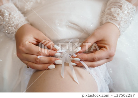 Elegant Bride Tying White Ribbon Garter. Wedding Day Close-up. Pearl Embellished Dress. Romantic. Soft Focus. Light Ivory Textures. Elegant Bride Tying White Ribbon Garter. Wedding Day Close-up. Pearl Embellished Dress. Romantic. Soft Focus. Light Ivory Textures. 127502287