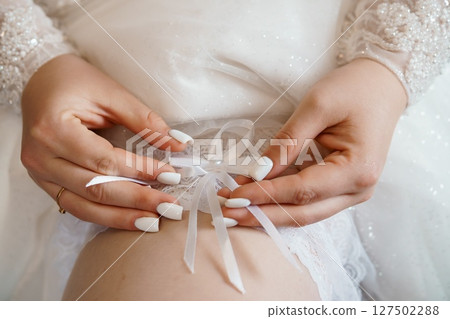 Close-up of Bride's Hands Tying White Ribbon, Wedding Garter Detail, Delicate Lace, Pearl Accents, Soft Focus, Romantic Bridal Scene. Close-up of Bride's Hands Tying White Ribbon, Wedding Garter Detail, Delicate Lace, Pearl Accents, Soft Focus, Romantic Bridal Scene. 127502288
