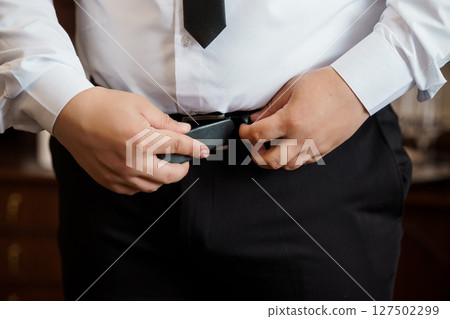 Man Fastening Black Belt With Suit and Tie, Formal Attire Preparation, Grooming Detail, Dark Clothing, Business Style, Studio Shot. 127502299