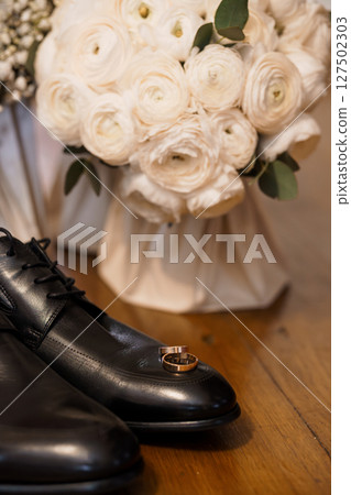 Wedding Day Details Still Life: Black Leather Shoes, Gold Rings, White Ranunculus Bouquet, Dark Wood Surface, Elegant Romantic Style 127502303