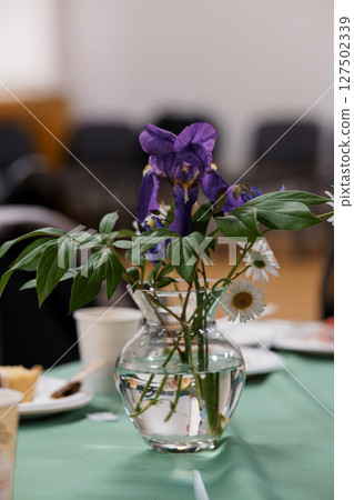Rustic Floral Arrangement in Glass Vase with Purple Delphiniums and Daisies on Green Tablecloth, Cozy Indoor Scene Rustic Floral Arrangement in Glass Vase with Purple Delphiniums and Daisies on Green Tablecloth, Cozy Indoor Scene 127502339