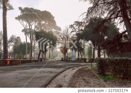 Serene scene of tranquil park surrounded by tall trees and prominent statue. Soft morning light filters through leaves, creating peaceful atmosphere. Villa Borghese gardens, Rome, Italy 127503174