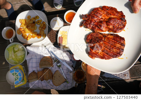 Two freshly grilled pork loin steaks on white plate held by hand over garden table, with more food from grill. Two freshly grilled pork loin steaks on white plate held by hand over garden table, with more food from grill. 127503440