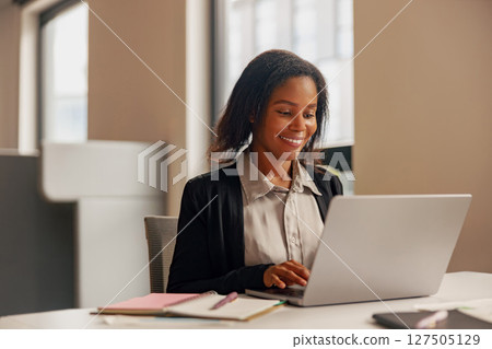 A cheerful professional is diligently working on their laptop in a modern office environment A cheerful professional is diligently working on their laptop in a modern office environment 127505129
