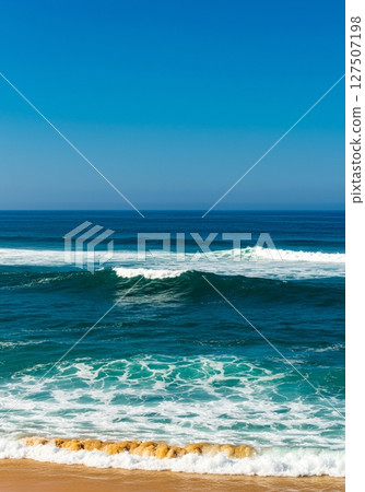 Ocean Waves Crashing on Sandy Beach Under Blue Sky 127507198