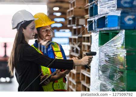 women in professional attire and man in safety gear collaborate on inventory tasks, scanning barcodes on pallets while surrounded by organized stacks of goods in the warehouse. 127509792