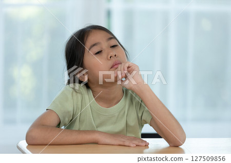 Young Girl in a Green Shirt Deep in Thought While Sitting at a Table in a Bright Indoor Environment 127509856