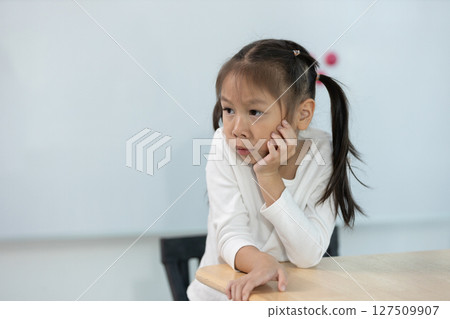 Young girl with long hair tied in ponytails rests her chin on her hand, displaying thoughtful expression as she sits at desk in bright classroom setting. Young girl with long hair tied in ponytails rests her chin on her hand, displaying thoughtful expression as she sits at desk in bright classroom setting. 127509907