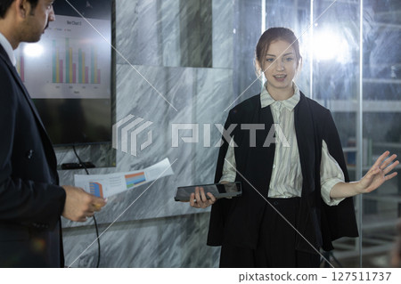Women in black suit confidently presents data analysis using tablet in sleek conference room. Colleagues attentively listen and engage with the information shared. 127511737