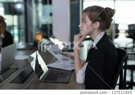 Professional woman is focused on her laptop holding document paper in office. Natural light is streaming through nearby windows, highlighting her concentration on work. Professional woman is focused on her laptop holding document paper in office. Natural light is streaming through nearby windows, highlighting her concentration on work. 127511954