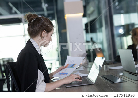 Professional woman is focused on her laptop holding document paper in office. Natural light is streaming through nearby windows, highlighting her concentration on work. 127511956
