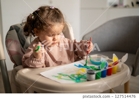 Side view of a young girl using a paintbrush with gouache while seated at a small art table. Childhood creativity in action. 127514476