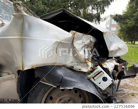 Severely damaged car after crash, showing twisted metal, rust, and broken parts. The vehicle appears abandoned near trees, emphasizing the impact of the collision and road hazards Severely damaged car after crash, showing twisted metal, rust, and broken parts. The vehicle appears abandoned near trees, emphasizing the impact of the collision and road hazards 127516896