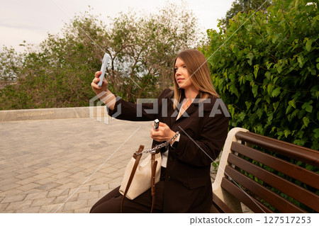 Young woman takes selfie seated on park bench outdoors. 127517253