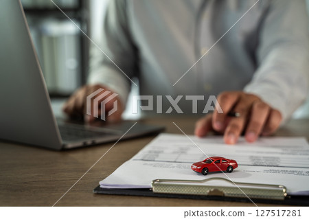 Insurance on paperwork assurance person is working desk with laptop, pointing document with small red car model top, indicating discussion about Insurance on paperwork assurance person is working desk with laptop, pointing document with small red car model top, indicating discussion about 127517281