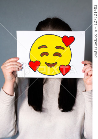 Woman holding smiling emoji mask with three hearts in front of face. Social media trends, Gen Z marketing, World emoji day 17 July. 127521402
