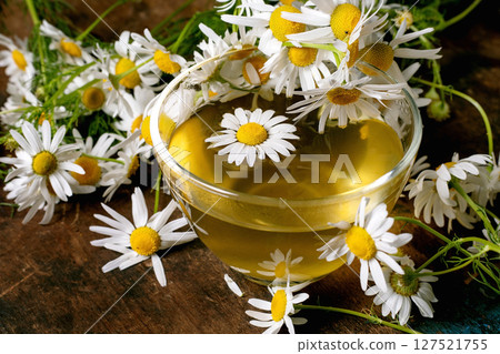 Glass cup chamomile tea surrounded by fresh white daisy flowers on wooden background. Herbal drink with natural blooms creates peaceful wellness scene. Glass cup chamomile tea surrounded by fresh white daisy flowers on wooden background. Herbal drink with natural blooms creates peaceful wellness scene. 127521755