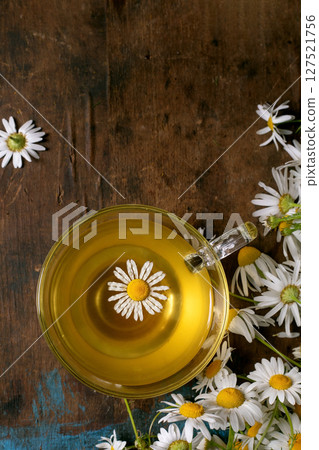 Glass cup chamomile tea with floating daisy flower surrounded by fresh chamomile flowers on rustic wooden background. Herbal tea concept natural remedy wellness. Glass cup chamomile tea with floating daisy flower surrounded by fresh chamomile flowers on rustic wooden background. Herbal tea concept natural remedy wellness. 127521756