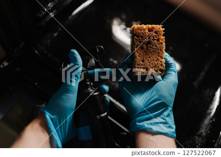 High-angle view of housewife in gloves spraying detergent on sponge, preparing to clean glass door of modern oven in kitchen with dark interior. Concept of cleanliness during routine chores. High-angle view of housewife in gloves spraying detergent on sponge, preparing to clean glass door of modern oven in kitchen with dark interior. Concept of cleanliness during routine chores. 127522067