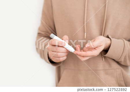 Close-up of a person using a lancet device to check blood sugar levels, beige hoodie against a white background.  127522892