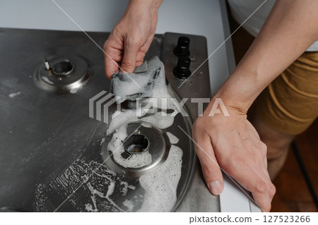 Close-up top view of housekeeper male hands scrubbing gas stove with detergent and rag during hygiene and maintenance in kitchen environments. Concept of domestic work, housekeeping and household. Close-up top view of housekeeper male hands scrubbing gas stove with detergent and rag during hygiene and maintenance in kitchen environments. Concept of domestic work, housekeeping and household. 127523266
