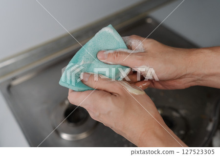 Top view of hand squeezing soapy water from sponge during cleaning dirty gas stove, to removing grime and grease, promoting hygiene and cleanliness in kitchen. Concept of domestic work and household. Top view of hand squeezing soapy water from sponge during cleaning dirty gas stove, to removing grime and grease, promoting hygiene and cleanliness in kitchen. Concept of domestic work and household. 127523305