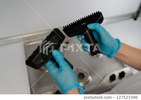 Top view of professional cleaner in rubber gloves holding and comparing two different brushes for steam cleaning dirty stainless steel stovetop, evaluating options for effective appliance maintenance. 127523306