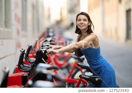 Brunette in blue dress rents electric bike on street of european city 127523543
