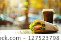 Paper coffee cup and fresh sandwich placed on an outdoor table with urban background and bokeh lights in the distance.  127523798