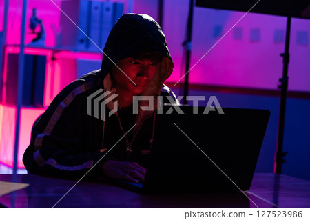 Young Man in Hoodie Working on Laptop in Dark Tech Environment Young Man in Hoodie Working on Laptop in Dark Tech Environment 127523986