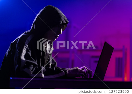 Young Man in Hoodie Working on Laptop in Dark Tech Environment Young Man in Hoodie Working on Laptop in Dark Tech Environment 127523990