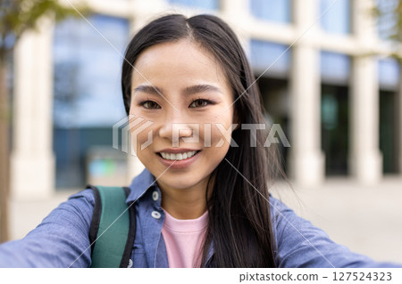 A smiling Asian woman takes a selfie, her face close to the camera. A backpack is visible, with a building in the background. 127524323
