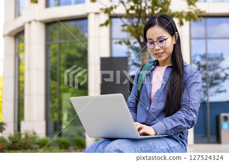 A young Asian woman uses a laptop outdoors, with a building and greenery in the background. 127524324