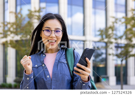 An excited Asian woman wearing glasses holds a phone and celebrates with her fist raised outside near a building. 127524326