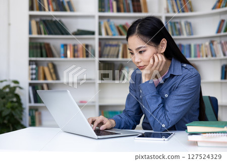 A focused Asian woman works on a laptop in a library, surrounded by books and a digital tablet. A focused Asian woman works on a laptop in a library, surrounded by books and a digital tablet. 127524329