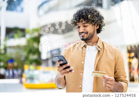 A smiling man in a shopping mall, holding a phone and a credit card, ready to make a purchase, depicting the convenience of online shopping. 127524458