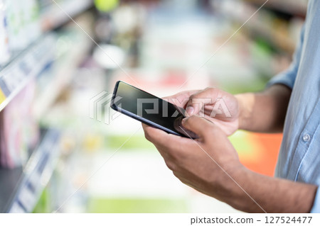 A person holding a smartphone in a supermarket aisle, possibly shopping or comparing prices. The soft focus highlights the technology and the setting. A person holding a smartphone in a supermarket aisle, possibly shopping or comparing prices. The soft focus highlights the technology and the setting. 127524477