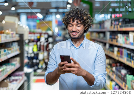 A smiling man uses his smartphone while browsing the aisles of a grocery store, surrounded by various products on shelves. 127524508