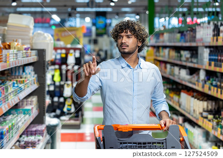 A man shops in a grocery store, extending his finger as he looks toward the shelves, pushing a shopping cart filled with items. 127524509