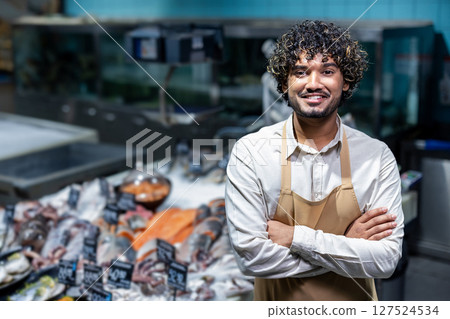 A smiling fishmonger stands confidently in front of a display of fresh seafood, ready to assist customers. He wears an apron. 127524534